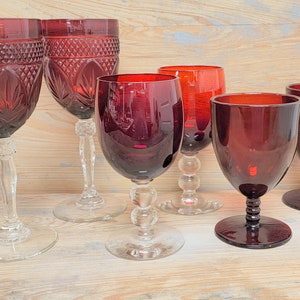 Peut inclure: Ensemble de six verres à vin rouge rubis. Les verres présentent des pieds et des bases transparents, avec des motifs de cuvette variés. Certains ont des motifs taillés complexes, d'autres une forme lisse et arrondie. Ils sont posés sur une surface en bois clair.