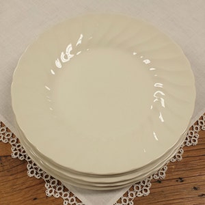 Peut inclure: Une pile de quatre assiettes en c&eacute;ramique blanc cass&eacute; avec un motif en spirale sur les bords. Les assiettes sont empil&eacute;es sur un napperon en dentelle blanche, cr&eacute;ant une esth&eacute;tique classique et &eacute;l&eacute;gante. Les assiettes sont sur une surface en bois.