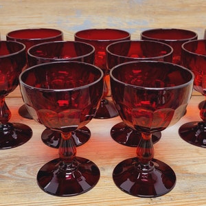 May include: A collection of ten ruby red wine glasses. Each glass features a round bowl with a faceted design, a slender stem, and a circular base. The glasses are arranged on a light-colored wooden surface.