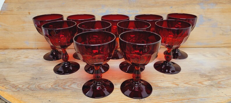 May include: A collection of ten ruby red wine glasses. Each glass features a round bowl with a faceted design, a slender stem, and a circular base. The glasses are arranged on a light-colored wooden surface.