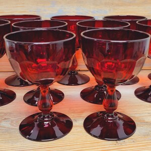 May include: A collection of ruby red wine glasses. Each glass has a round base, a decorative stem, and a bowl-shaped top. The glasses are arranged on a light-colored wooden surface, creating a visually appealing display.