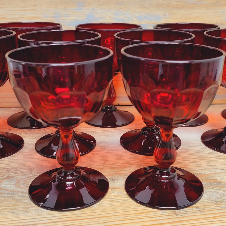 May include: A collection of ruby red wine glasses. Each glass has a round base, a decorative stem, and a bowl-shaped top. The glasses are arranged on a light-colored wooden surface, creating a visually appealing display.