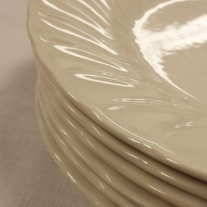 Peut inclure: Une pile d'assiettes en c&eacute;ramique blanc cass&eacute;. Chaque assiette pr&eacute;sente un motif de bordure cannel&eacute; et tourbillonnant. Les assiettes sont empil&eacute;es, la premi&egrave;re &eacute;tant mise au point.