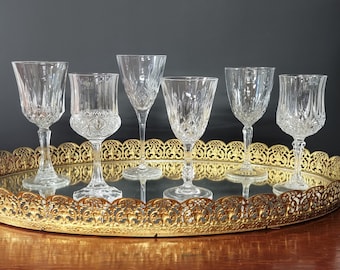 May include: Six clear glass goblets with intricate cut designs sit on a gold mirrored tray with a decorative scalloped edge.
