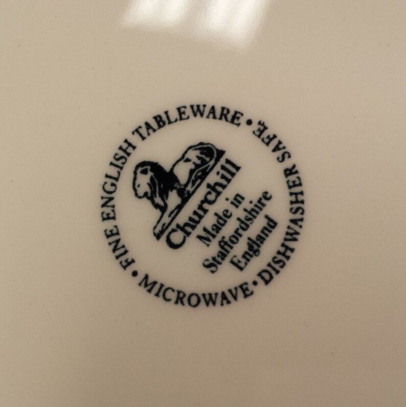 Peut inclure: Gros plan sur une assiette cr&egrave;me avec un logo circulaire. Le logo pr&eacute;sente le mot "Churchill" et un embl&egrave;me de lion, avec du texte indiquant qu'il s'agit de fine vaisselle anglaise, allant au micro-ondes et au lave-vaisselle, fabriqu&eacute;e en Angleterre.
