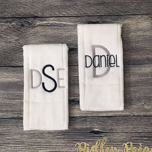 May include: Two white burp cloths with embroidered names. The first cloth has the name "DSE" embroidered in gray and black thread. The second cloth has the name "Daniel" embroidered in gray and black thread.