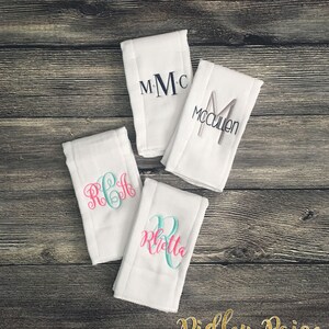 May include: Four white burp cloths with different embroidered names in pink, teal, and gray. The names are "MMC", "McCullen", "RGH", and "Kietta".