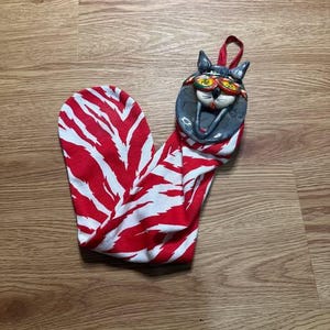 May include: A whimsical Christmas stocking featuring a red and white zebra stripe pattern. The top of the stocking has a ceramic cat face with sunglasses and the word "flat" on the glasses. A red loop is attached for hanging.