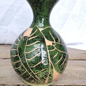 May include: A green ceramic vase with a speckled glaze and a leaf design. The vase has a round body and a narrow neck.