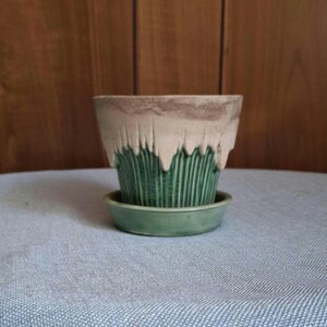 May include: A decorative ceramic plant pot with a distinctive design. The top section is a light beige with a textured, dripped effect, leading to a green, vertically-lined lower part. It rests on a matching green saucer.