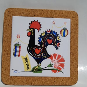 May include: A cork-backed trivet with a white ceramic tile featuring a black rooster with red, yellow, and blue accents. The rooster is standing on a branch with a red flower and the word "PORTUGAL" written in yellow. The tile also features two red, white, and blue lanterns.