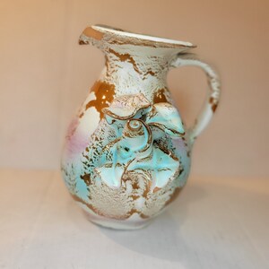 May include: A ceramic jug with a handle and a spout. The jug is decorated with a distressed white, gold, and turquoise finish. A floral design is on the front. This decorative jug is suitable for display or as a decorative item.