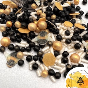 May include: Black and gold bee-themed sprinkles in a clear jar. The sprinkles include black and gold balls, white and gold rods, and black and gold bee shapes. The jar is spilling out the sprinkles onto a white surface. The image includes the text "Bee Box Design Studio".