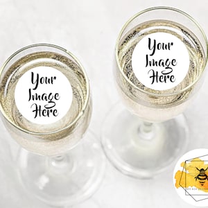 May include: Two champagne flutes filled with a sparkling beverage. Each glass has a white circular sticker with the text "Your Image Here" in black script. The glasses are on a white surface.