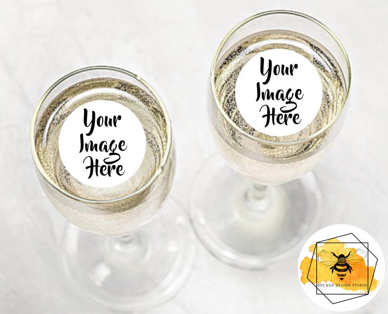 May include: Two champagne flutes filled with a sparkling beverage. Each glass has a white circular sticker with the text "Your Image Here" in black script. The glasses are on a white surface.