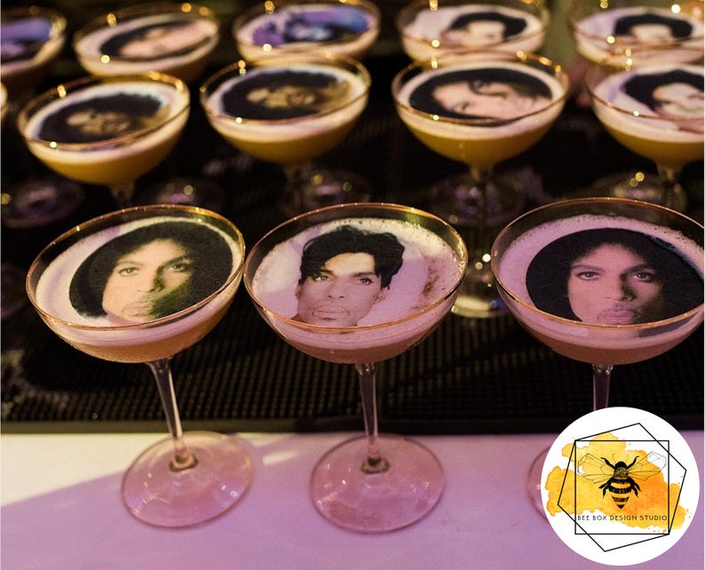 May include: Cocktails with a foamy top and a gold rim, featuring a portrait of a musician. The drinks are served in stemmed glasses. The image is taken from a low angle, with a blurred background.