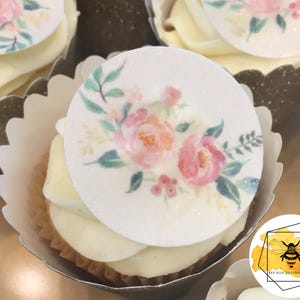K&ouml;nnte beinhalten: Ein Cupcake mit wei&szlig;em Frosting und einem runden essbaren Bild von rosa und gr&uuml;nen Aquarellblumen. Der Cupcake befindet sich in einer wei&szlig;en Papierform mit einem gewellten Rand.