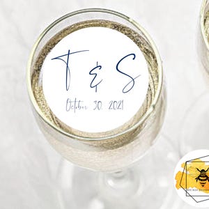 May include: White round sticker with the text "T & S" in blue cursive font and the date "October 30, 2021" printed below. The sticker is attached to a champagne flute.