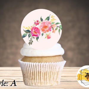 K&ouml;nnte beinhalten: Ein Cupcake mit einem runden, wei&szlig;en Frosting-Oberteil und einem essbaren Topper mit pinkfarbenem und gelbem Blumendesign. Der Cupcake steht auf einer Holzoberfl&auml;che. Der essbare Topper tr&auml;gt den Text "Style: A".