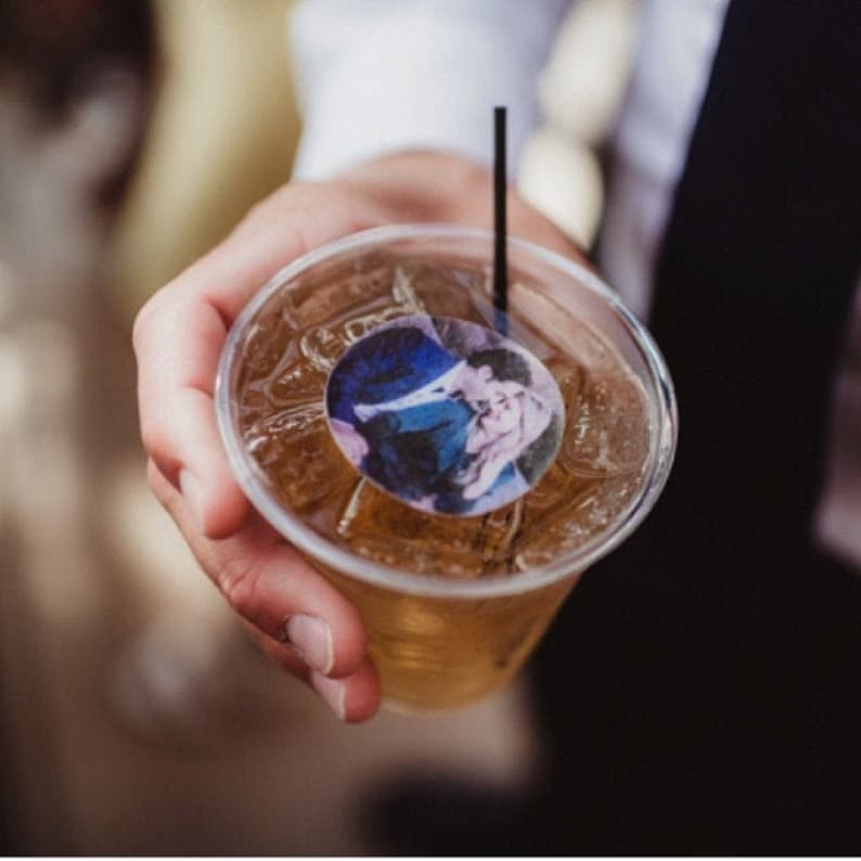 May include: A clear plastic cup filled with ice and a light brown liquid, possibly a cocktail. A black straw and a circular sticker with a blue and purple design are visible. The cup is held by a person.