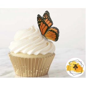 Puede incluir: Un cupcake con glaseado blanco y una decoración de mariposa monarca. El cupcake está en un envoltorio de papel dorado. La mariposa tiene alas naranjas y negras. La imagen incluye un logotipo con una abeja.
