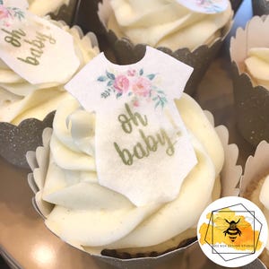 K&ouml;nnte beinhalten: Nahaufnahme eines Cupcakes mit wei&szlig;em Frosting und einem wei&szlig;en Fondant-Topper in Form eines Stramplers mit floralen Akzenten. Der Topper tr&auml;gt den Text "oh baby!" in goldenen Buchstaben.