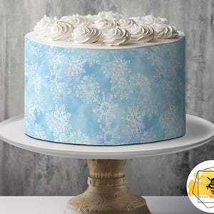 Könnte beinhalten: Ein blauer und weißer Kuchen, der mit weißem Frosting und einem Schneeflockenmuster verziert ist. Der Kuchen steht auf einem weißen Kuchenständer mit einem Holzsockel.