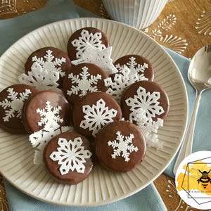 May include: A plate of chocolate biscuits decorated with white snowflake designs. The biscuits are arranged in a circle on a white plate with a blue napkin and a silver spoon in the background.