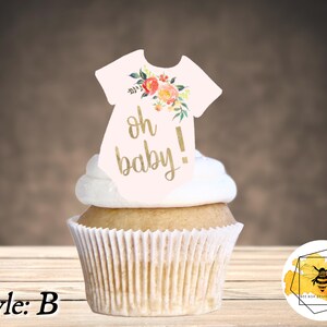 K&ouml;nnte beinhalten: Ein Cupcake mit wei&szlig;em Frosting und einem rosa-wei&szlig;en Baby-Strampler-Topper mit goldener Aufschrift "oh baby!" und floralen Akzenten.