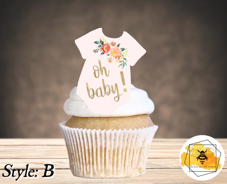 K&ouml;nnte beinhalten: Ein Cupcake mit wei&szlig;em Frosting und einem rosa-wei&szlig;en Baby-Strampler-Topper mit goldener Aufschrift "oh baby!" und floralen Akzenten.