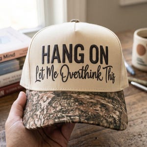 May include: A cream-colored baseball cap with a camouflage-patterned brim. The front panel features the text "HANG ON" in bold, dark block letters above the script text "Let Me Overthink This" in a matching dark color.