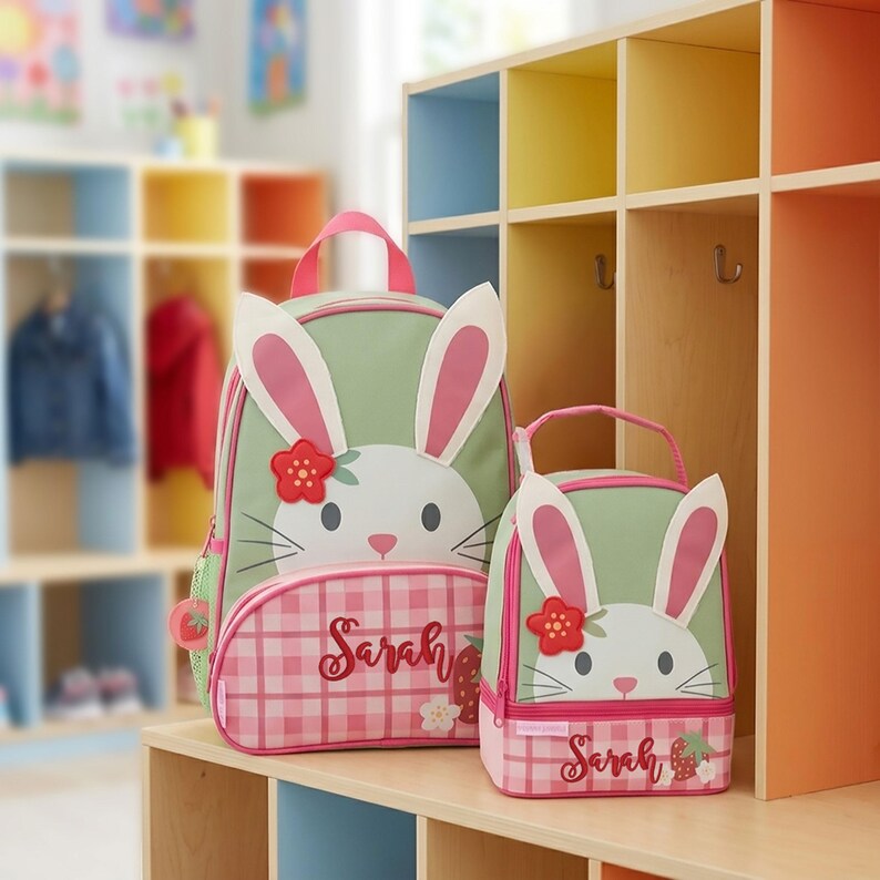 May include: A sage green and pink plaid backpack and matching lunch bag set featuring a white bunny face with long ears, a red flower accent, and the name Sarah embroidered in cursive script on the front pocket of each item.