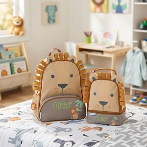 May include: A tan and gray lion-themed backpack and matching lunch bag set. Both items feature a lion face with a mane, a gray bottom panel with the name Oliver embroidered in green, and a small lion graphic next to the name on each piece.