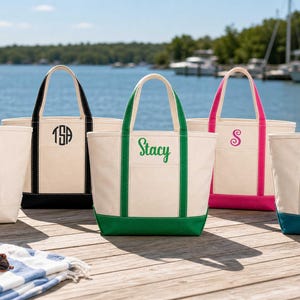 May include: Five off-white canvas tote bags with colored handles and bases. Each bag features a unique embroidered monogram or name: LSA, TSA, Stacy, S, and Taylor. The bags are arranged in a row on a wooden dock near a body of water.