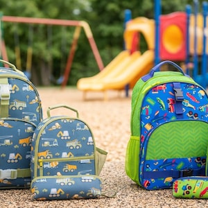 May include: Two sets of backpacks, lunch bags, and pencil cases. One set features a blue background with yellow construction vehicles. The other set features a blue background with colorful vehicles, green accents, and a chevron pattern on the pockets.