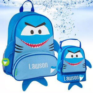 May include: A blue shark-themed backpack and matching lunch bag set. Both items feature a cartoon shark face with white eyes, black eyebrows, and a red mouth with sharp teeth. The name Lawson is printed in white on the front of both items.
