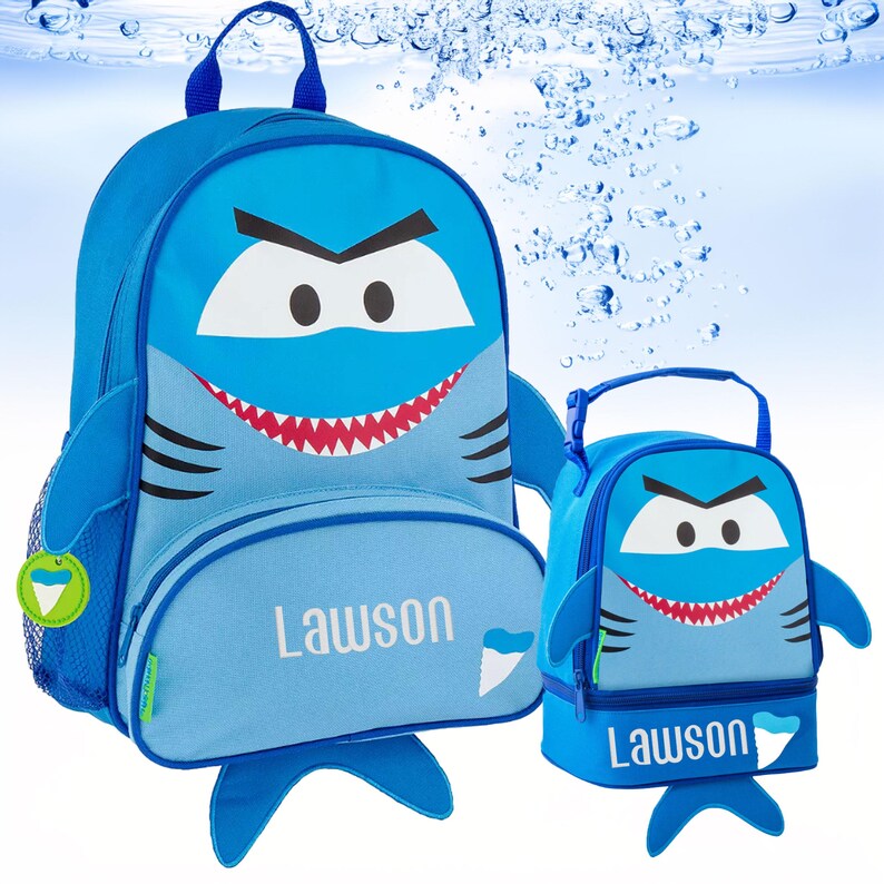 May include: A blue shark-themed backpack and matching lunch bag set. Both items feature a cartoon shark face with white eyes, black eyebrows, and a red mouth with sharp teeth. The name Lawson is printed in white on the front of both items.