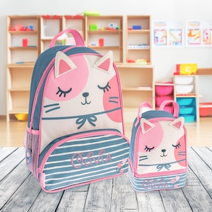 May include: A blue and white backpack and matching lunch bag set featuring a cat face design with pink accents and closed eyes. Both items are personalized with the name Olivia in pink script text and include small pink tulip illustrations.