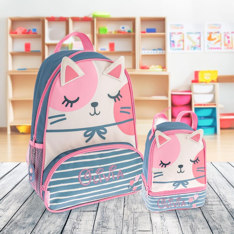 May include: A blue and white backpack and matching lunch bag set featuring a cat face design with pink accents and closed eyes. Both items are personalized with the name Olivia in pink script text and include small pink tulip illustrations.