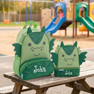May include: A light green dragon-themed backpack and matching lunch bag set. Both items feature dark green dragon wings, scale patterns, and the name John embroidered in white on the front pocket. The backpack has side mesh pockets.