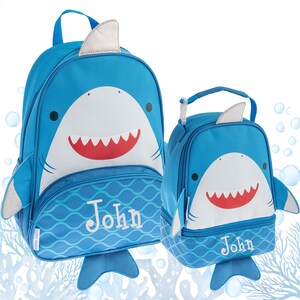May include: A blue shark-themed backpack and matching lunch bag set, both featuring a white shark face with a red mouth, silver fins, and a blue wave pattern at the base. Each item is personalized with the name John embroidered in white text.