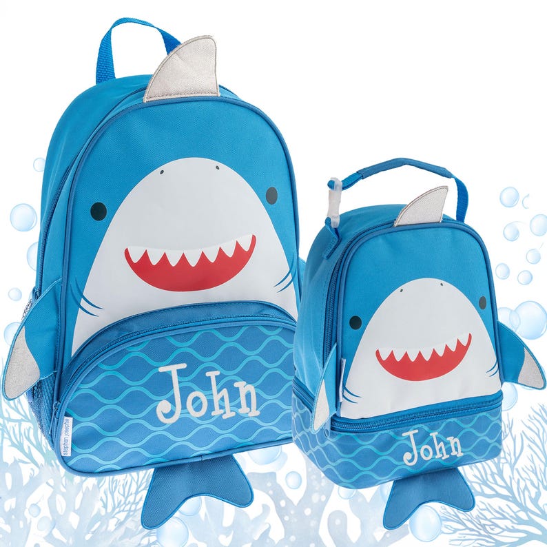 May include: A blue shark-themed backpack and matching lunch bag set, both featuring a white shark face with a red mouth, silver fins, and a blue wave pattern at the base. Each item is personalized with the name John embroidered in white text.