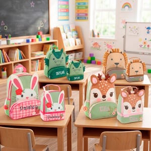 May include: A collection of animal-themed backpacks and matching lunch bags featuring embroidered names including Paisley, Skylar, and Ella. The designs include a green rabbit, a green dragon, a tan lion, and a brown deer, all displayed on wooden desks.