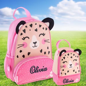 May include: A pink backpack and a matching smaller lunch bag, both featuring a cartoon leopard face with black heart-shaped spots, pink cheeks, and black ears. The front pocket of each item is personalized with the name Olivia in black script.