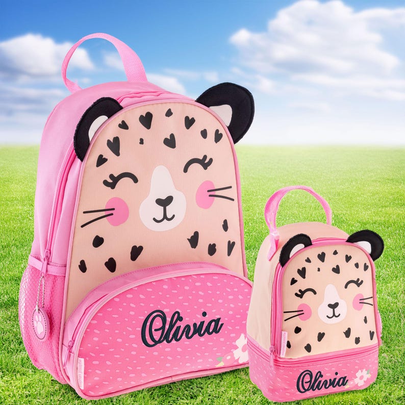 May include: A pink backpack and a matching smaller lunch bag, both featuring a cartoon leopard face with black heart-shaped spots, pink cheeks, and black ears. The front pocket of each item is personalized with the name Olivia in black script.