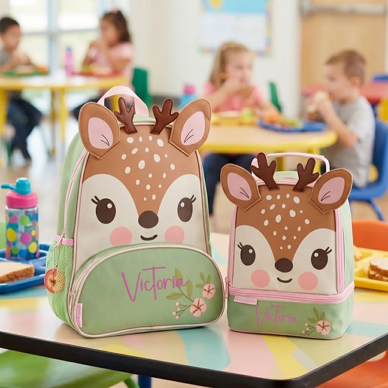 May include: A sage green backpack and a matching lunch bag, both featuring a brown deer face design with antlers, white spots, and pink cheeks. The name Victoria is printed in pink script on the front of both items, accompanied by a small floral graphic.