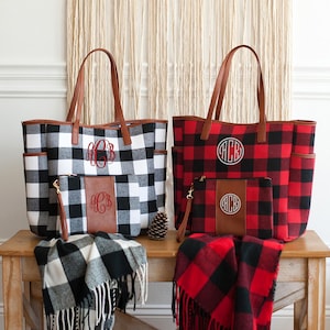 May include: Two large tote bags with matching smaller zippered pouches. The tote bags are made of a red and black buffalo plaid fabric and a black and white buffalo plaid fabric. Both bags have brown leather handles and a brown leather patch with a monogram on the front. The bags are sitting on a wooden table with matching buffalo plaid blankets.