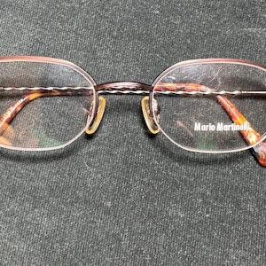May include: Oval-shaped eyeglasses with a brown metal frame and clear lenses. The temples have a tortoiseshell pattern and the brand name "Mario Martinelli" is visible on the lens. The glasses are set against a dark background.