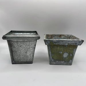May include: Two square metal planters with a tapered shape. The left planter has a bright, uniform galvanized silver finish. The right planter features a weathered, distressed appearance with patches of green oxidation and brown rust on the surface.