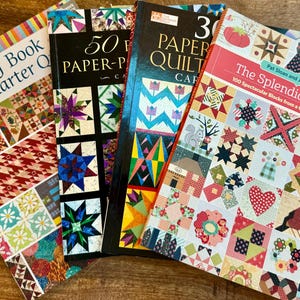 Puede incluir: Cuatro libros de acolchado con portadas coloridas que presentan diseños de patchwork. Los títulos son "The Big Book of Fat-Quarter Quilts", "50 Easy Paper-Pieced Quilts", "30 Paper-Pieced Quilts" y "The Splendid Sampler: 100 Spectacular Blocks from a Community of Quilters".